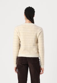 Krémový svetr s texturovaným kostkovaným vzorem, s kulatým výstřihem a žebrovanými manžetami. Spárovaný s hnědými sametovými kalhotami.