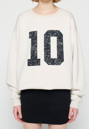 Person trägt ein übergroßes hellbeiges Sweatshirt mit einer abgenutzten schwarzen Zahl "10" und einen kurzen schwarzen Rock.