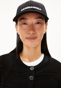 Schwarze Mütze mit weißer "ARMEDANGELS" Stickerei, kombiniert mit einem strukturierten schwarzen Cardigan mit zwei schwarzen Knöpfen und einem weißen Kragen.