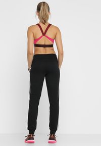 Zwarte sportbroek met witte strepen, gecombineerd met een bordeauxrode en roze sportbeha met een kruis-achter ontwerp. Schoenen zijn roze en bruin.