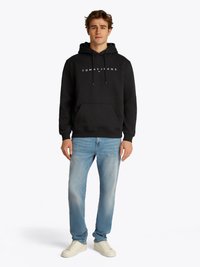 Svart hettegenser med lomme foran og hvit "TOMMY JEANS" logo, sammen med lyseblå denimjeans og hvite sneakers.
