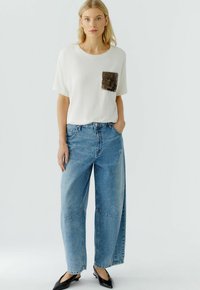 Witte katoenen t-shirt met een bruine pailletten zak, gecombineerd met lichtblauwe wijde jeans. Zwarte schoenen maken de outfit compleet. Eenvoudig ontwerp en casual pasvorm.