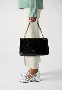 Black faux leather handbag with a textured, quilted pattern and gold chain handle; paired with silver heeled shoes and light-colored trousers.