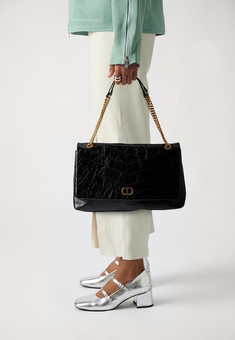 Black faux leather handbag with a textured, quilted pattern and gold chain handle; paired with silver heeled shoes and light-colored trousers.