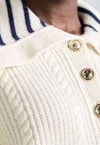 Gestrickter Pullover in Creme mit Zopfmuster, einem breiten Kragen mit marineblauen Streifen und goldenen Knöpfen an der vorderen Knopfleiste.