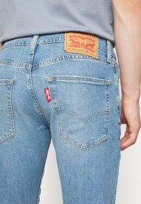 Jean en denim Levi’s bleu clair avec poches arrière, étiquette logo rouge et patch de marque en cuir à la taille, porté par une personne en chemise grise.