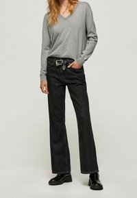 Person, der einen grauen V-Ausschnitt-Pullover, schwarze weit geschnittene Jeans mit einem schwarzen Gürtel und schwarze klobige Stiefeletten trägt, steht vor einem schlichten Hintergrund.