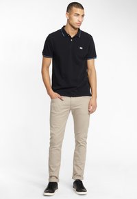 Polo noir avec des bordures bleues, pantalon beige et baskets noires. Le polo présente un petit logo sur la poitrine, en matière coton lisse.