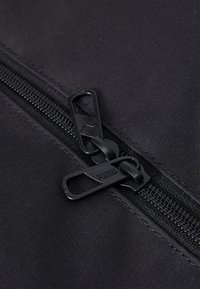 Zipper in tessuto nero con design a doppio cursore. Le linguette di apertura hanno una finitura opaca con un dettaglio del logo. I denti della zip sono visibili lungo il nastro.