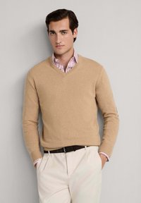 Maglione beige a collo a V realizzato in materiale morbido, con maniche lunghe. Indossato sopra una camicia a righe rosa chiaro e bianca con i polsini in vista.