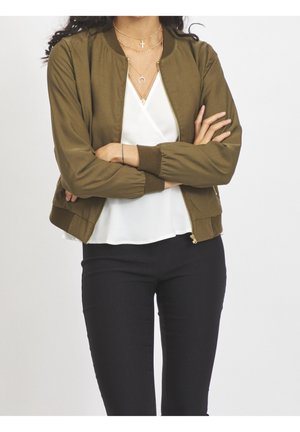 Personne, les bras croisés, portant une veste zippée vert olive, un chemisier blanc, un pantalon noir et des colliers dorés superposés.