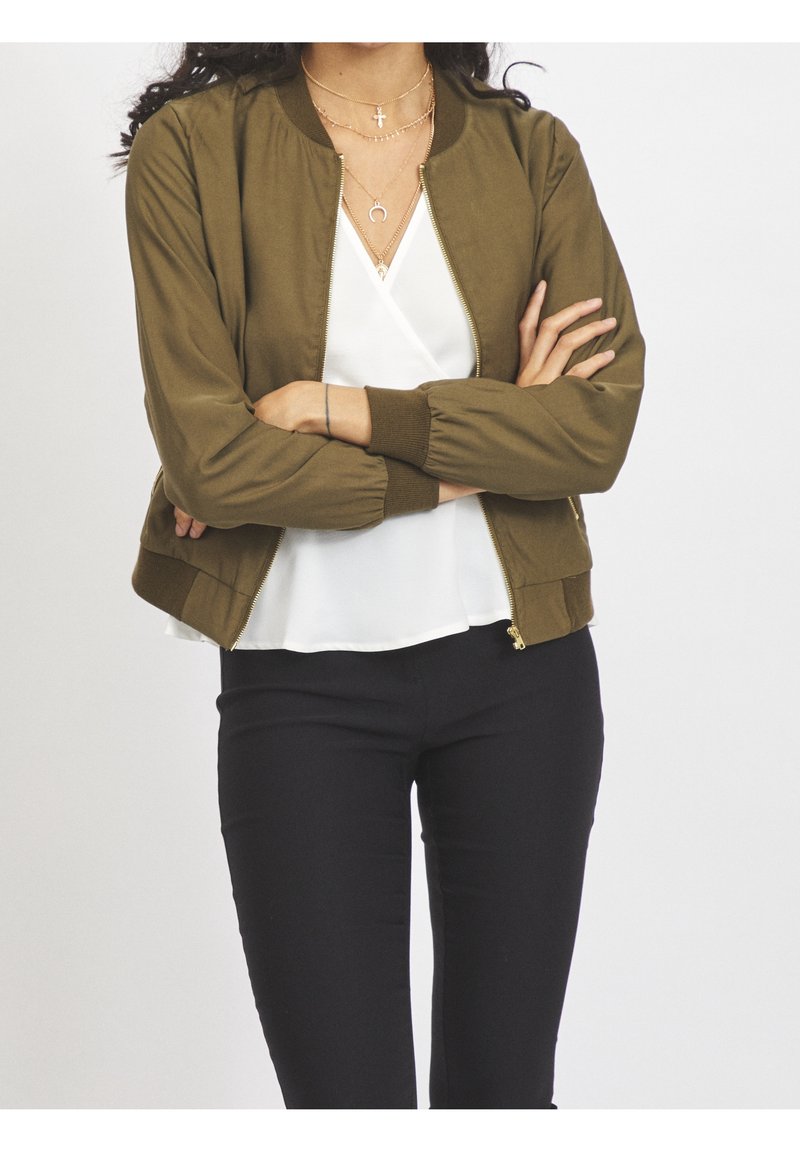 Personne, les bras croisés, portant une veste zippée vert olive, un chemisier blanc, un pantalon noir et des colliers dorés superposés.