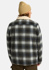 Een persoon in een beige beanie en een checkered flanellen jas met een witte, bontgevoerde kraag, gezien vanaf de achterkant.