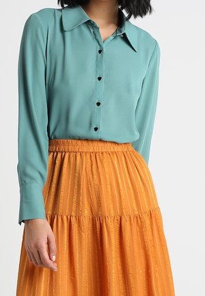 Frau trägt eine türkise Bluse mit langen Ärmeln und herzförmigen Knöpfen, die in einen hoch taillierten, strukturierten orangen Rock gesteckt ist.