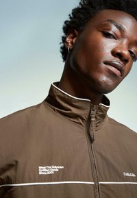 Brown zip-up jacket with high collar, white accents, and logo text. Features a textured surface and a sporty design.