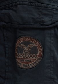 Mørkeblå stof med et rundt, broderet patch med vinger og ternede mønstre. Teksten inkluderer "International Speedway" og "Daytona."
