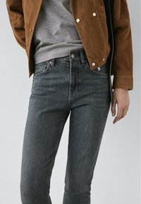 Personne portant un jean gris foncé, une chemise grise et une veste en daim marron, debout devant un fond uni gris clair.