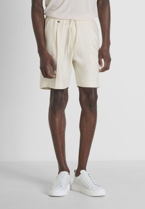 Pantalones cortos de lino en beige claro con cintura elástica y cordón, que presentan un corte relajado y bolsillos laterales. Zapatillas blancas completan el conjunto.