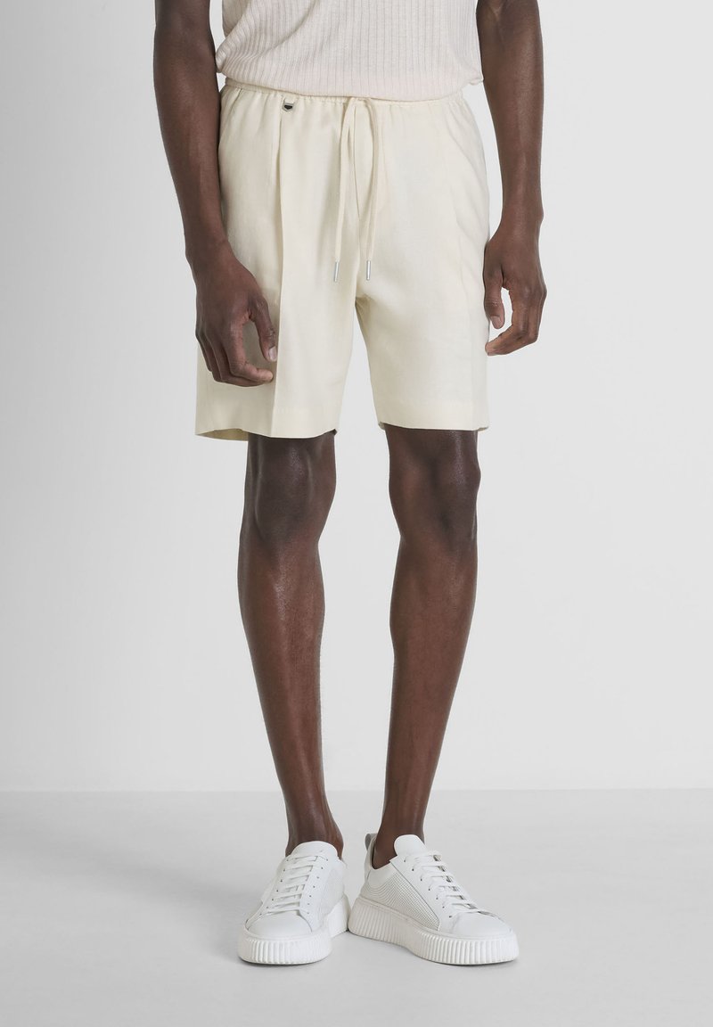 Bermuda in lino di un beige chiaro con elastico in vita e lacci, caratterizzati da una vestibilità morbida e tasche laterali. Sneakers bianche completano il look.