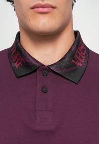 Homem vestido com uma camisa polo púrpura escura com colarinho preto, apresentando letras em bordô e padrões decorativos, dois botões pretos abotoados.
