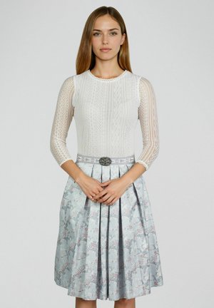 Junge Frau mit glattem braunem Haar trägt eine weiße Spitzenbluse und einen hellblauen, geblümten, plissierten Rock mit dekorativem Gürtel.