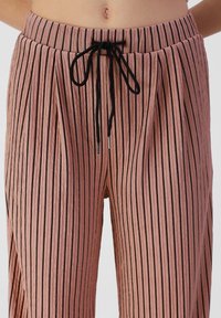 Záhybové, růžové kalhoty s vertikálními černými pruhy; zahrnují elastický pás a stahovací šňůrku pro nastavitelný střih.