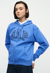 Νεαρό άτομο με κοντά σκουρόχρωμα μαλλιά, που φοράει μπλε φούτερ GAP πάνω από λευκό πουκάμισο, στέκεται με τα χέρια πίσω από την πλάτη μπροστά σε λευκό φόντο.