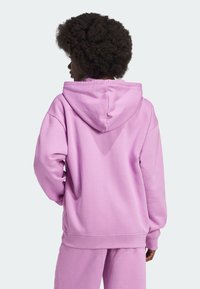 Osoba s kudrnatými vlasy, oblečená v bledě fialové mikině a odpovídajících kalhotách, otočená zády k jednobarevnému světlému pozadí.