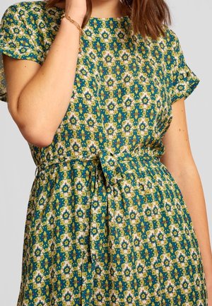 Femme portant une robe à motif floral turquoise et jaune, manches courtes, avec une ceinture nouée à la taille, visible des épaules aux hanches.