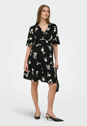 Kvinde iført en sort knælængde kjole med hvidt blomstertryk, der holder en sort håndtaske og bærer sorte lave hælslingbacks.