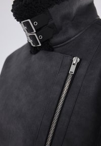 Czarna skórzana kurtka o teksturowanym wykończeniu, z wysokim kołnierzem, metalowymi klamrami i boczną kieszenią na zamek błyskawiczny ze srebrnym zamkiem.