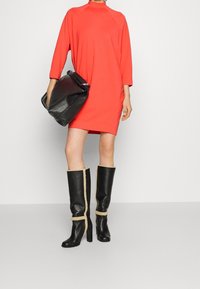 Rotes, übergroßes Kleid mit hohem Kragen, getragen mit knielangen schwarzen Lederstiefeln mit beigen Akzenten. Schwarze, lässige Tasche in einer Hand gehalten.