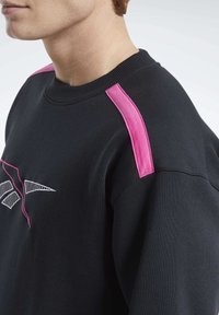 Acercamiento de una persona que lleva una sudadera negra con acentos rosas en los hombros y un logotipo geométrico parcialmente visible en el pecho.