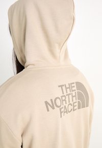 Sudadera beige hecha de material suave, con capucha y gran logo gris en la parte posterior. Se aprecian costuras en el escote.