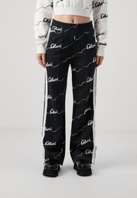 Pantalones negros con patrón de texto ondulado blanco, cinturilla elástica, paneles laterales y un corte relajado. El material parece suave y ligero.