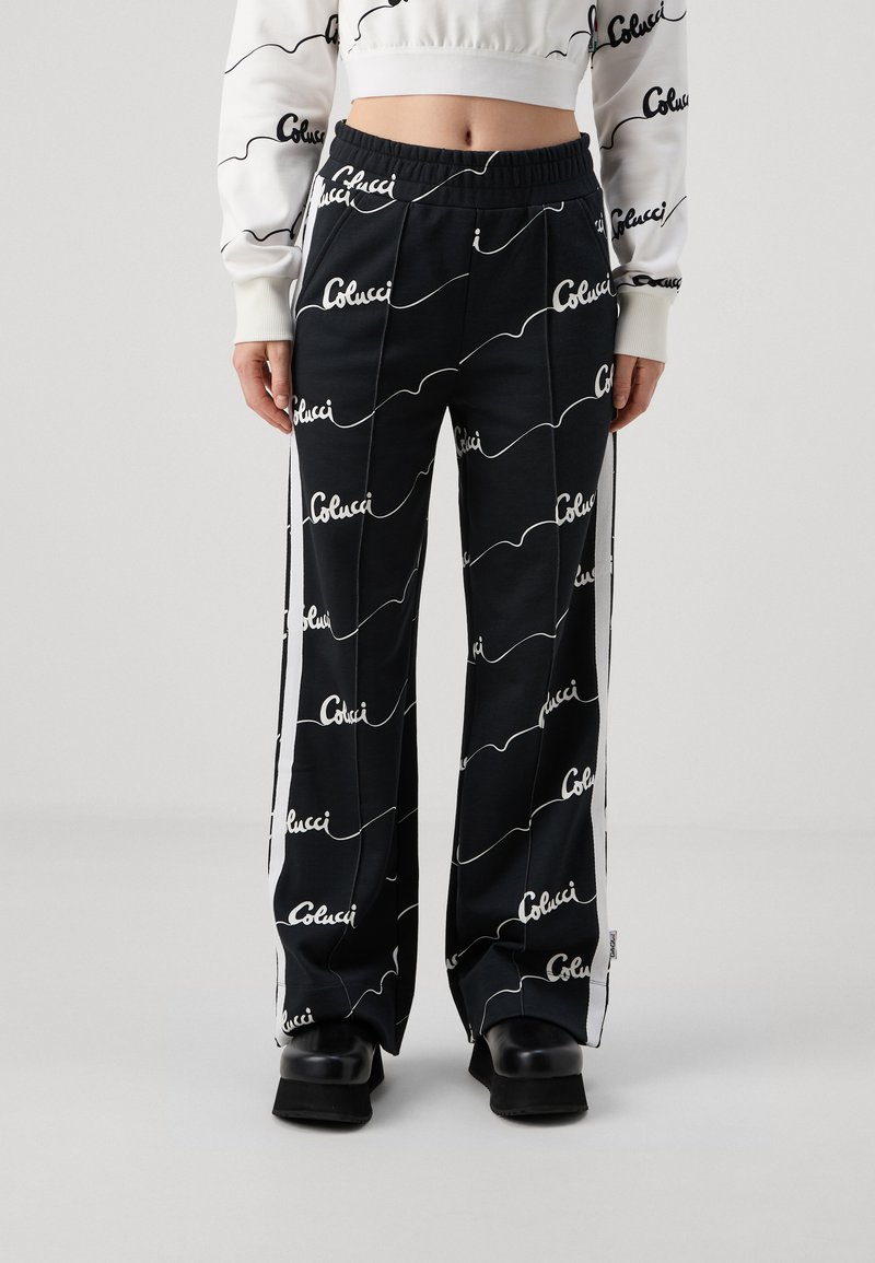 Pantalones negros con patrón de texto ondulado blanco, cinturilla elástica, paneles laterales y un corte relajado. El material parece suave y ligero.