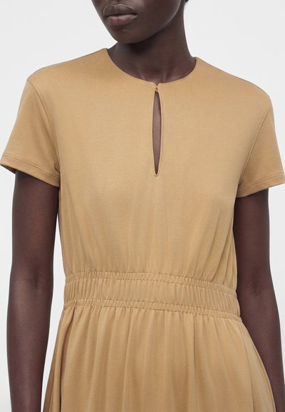 Gros plan d'une personne portant une robe beige à manches courtes avec une taille froncée et un petit détail d'encolure en forme de clé à l'avant.