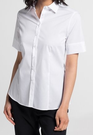 Woman wearing a fitted white short-sleeve button-up shirt with a collar and black pants against a plain background.