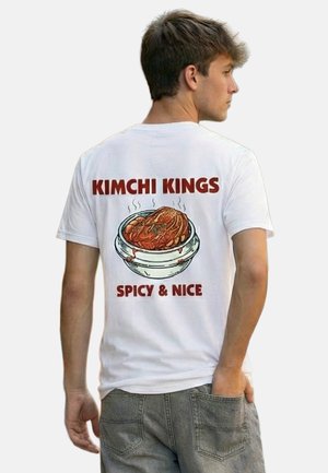 Junger Mann seitlich, trägt ein weißes Hemd mit „KIMCHI KINGS“ und einer dampfenden Schüssel Kimchi mit der Aufschrift „SPICY & NICE“ auf dem Rücken.