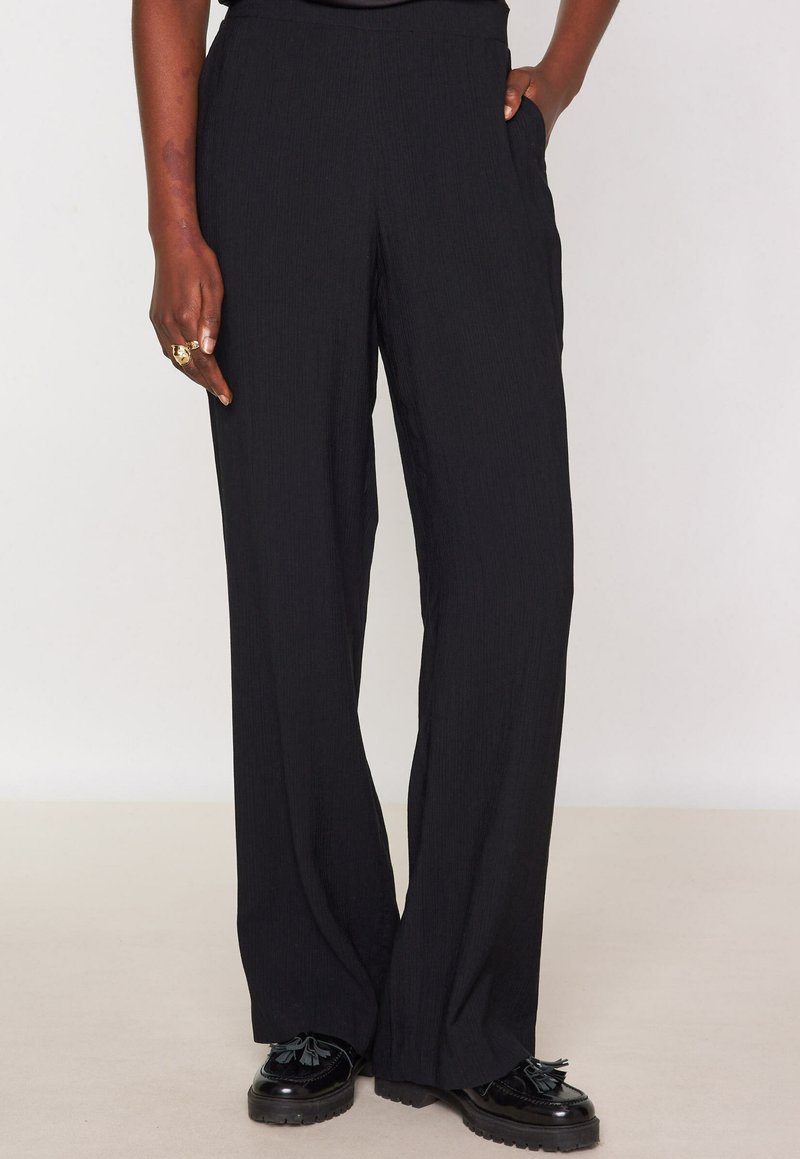 Vrouw in losse zwarte broek en zwarte tassel loafers, met een hand in haar zak en de andere aan haar zij met gouden ringen.