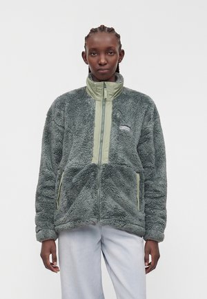 Person trägt eine grau-grüne Fleecejacke mit hohem Kragen, zwei Fronttaschen und einem teilweisen Frontreißverschluss.