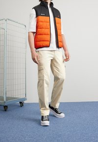 Veste matelassée rouge et noire portée sur un t-shirt blanc, associée à un pantalon cargo beige et des baskets noires. Texture lisse, motif matelassé sur la veste.