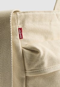 Bolsa de lona en beige con superficie texturizada, que presenta una etiqueta roja de Levi's y un detalle de bolsillo lateral cosido.
