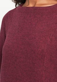 Bordo pleteni pulover s rebrastom teksturom, širokim izrezom i bešavnim ramenima, naglašavajući mekoću i toplinu.
