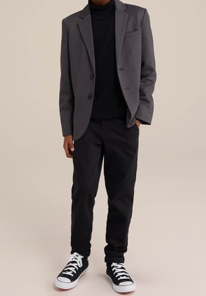 Blazer gris porté sur un col roulé noir, associé à un pantalon noir et des baskets noires. Le blazer comporte deux boutons et une poche poitrine.