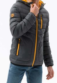 Chaqueta acolchada gris con capucha, que presenta detalles y cremalleras amarillas, textura acolchada y dos bolsillos con cremallera en la parte delantera.