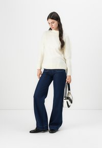Weißen Strickpullover mit hohem Kragen, kombiniert mit dunkelblauen Schlagjeans und schwarzen Schuhen. Hält eine schwarz-weiße Handtasche.