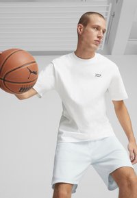 Biely bavlnený tričko s čiernym geometrickým logom, v kombinácii so svetlomodrými atletickými šortkami z oka. Osoba drží basketbal.