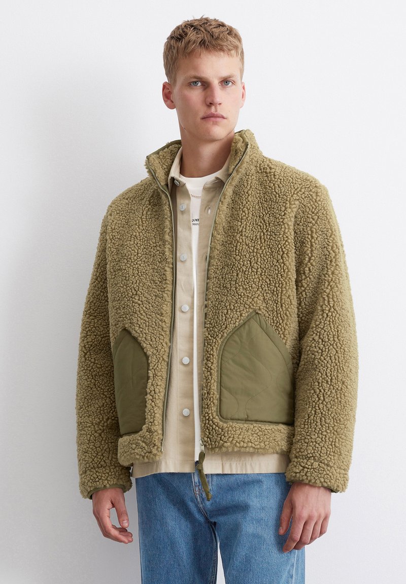 Khaki-Fleece-Jacke mit durchgehendem Reißverschluss, hohem Kragen und olivgrünen, gesteppten Taschen. Weicher Stoff und lockere Passform, getragen über einem cremfarbenen Hemd.