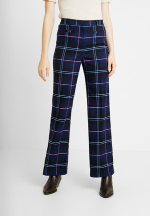 Femme portant un pantalon à carreaux bleu marine avec des rayures bleu clair et rose, associé à des bottines noires et un haut beige clair, debout sur fond blanc.
